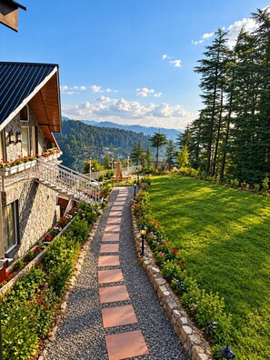 A pathway leading to a house with a stone facade and a balcony with red flowers.