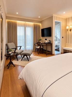 A hotel room with a large bed, a desk with a TV, a seating area with chairs, and a view of the city through the window. - La Foret - A Boutique Resort (Shimla)