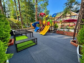 Un terrain de jeux avec des toboggans et des structures d’escalade, entouré d’arbres, et un bâtiment avec un toit rouge.