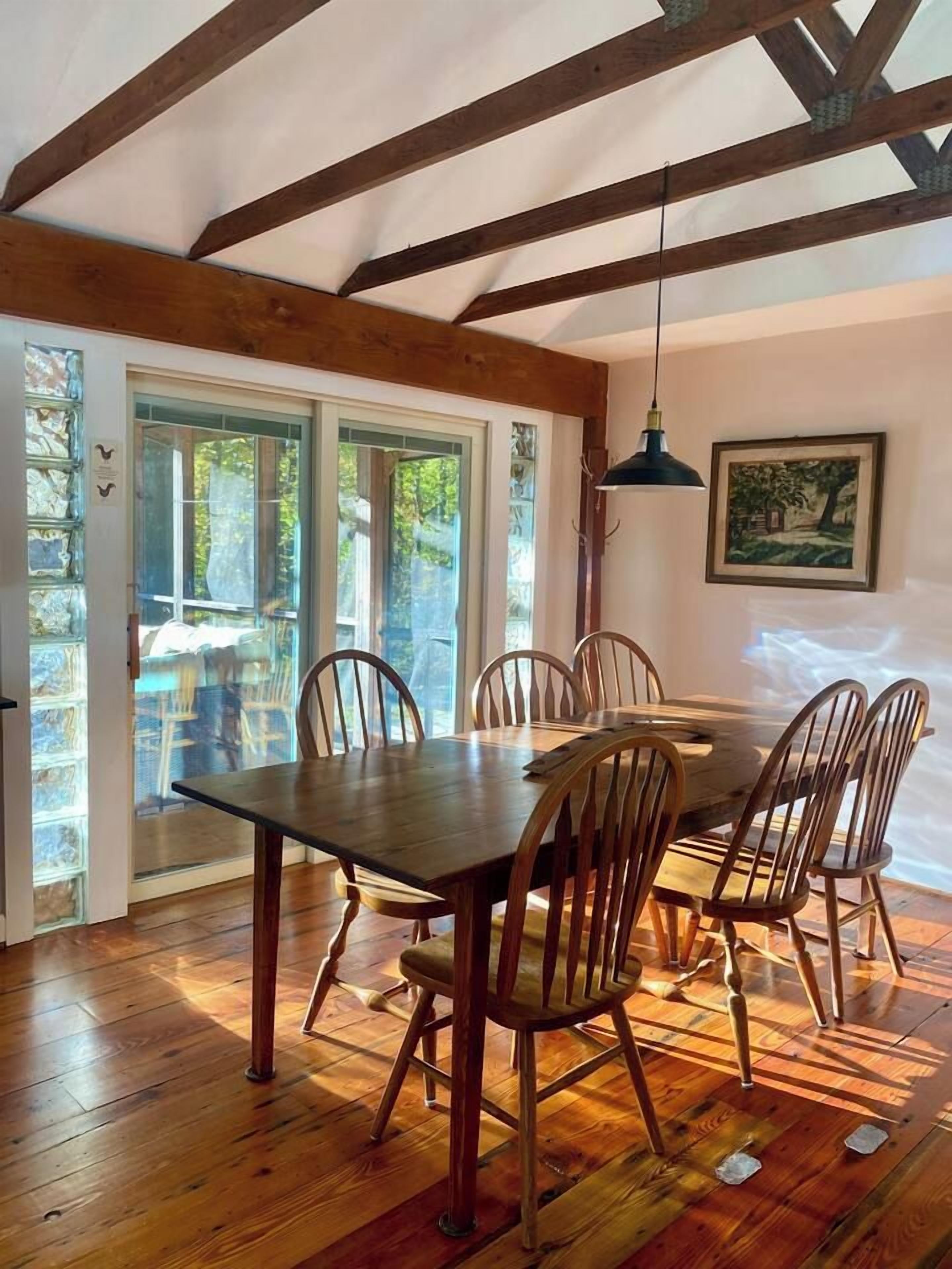 A dining area with a wooden table and chairs, a hanging light fixture, and a view through glass doors to an outdoor area.