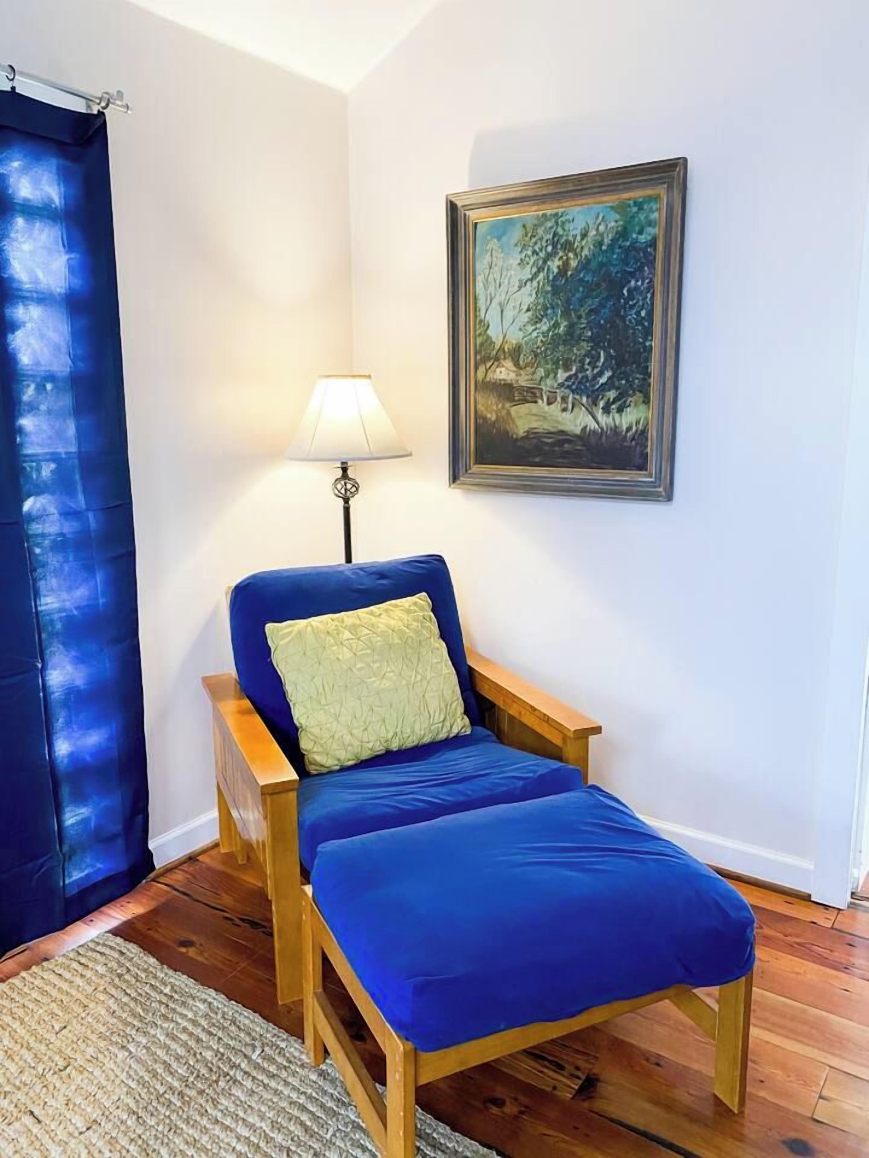 Ein Zimmer mit einem blauen Sofa, einem Holztischchen, einer Lampe und einem gerahmten Gemälde an der Wand.
