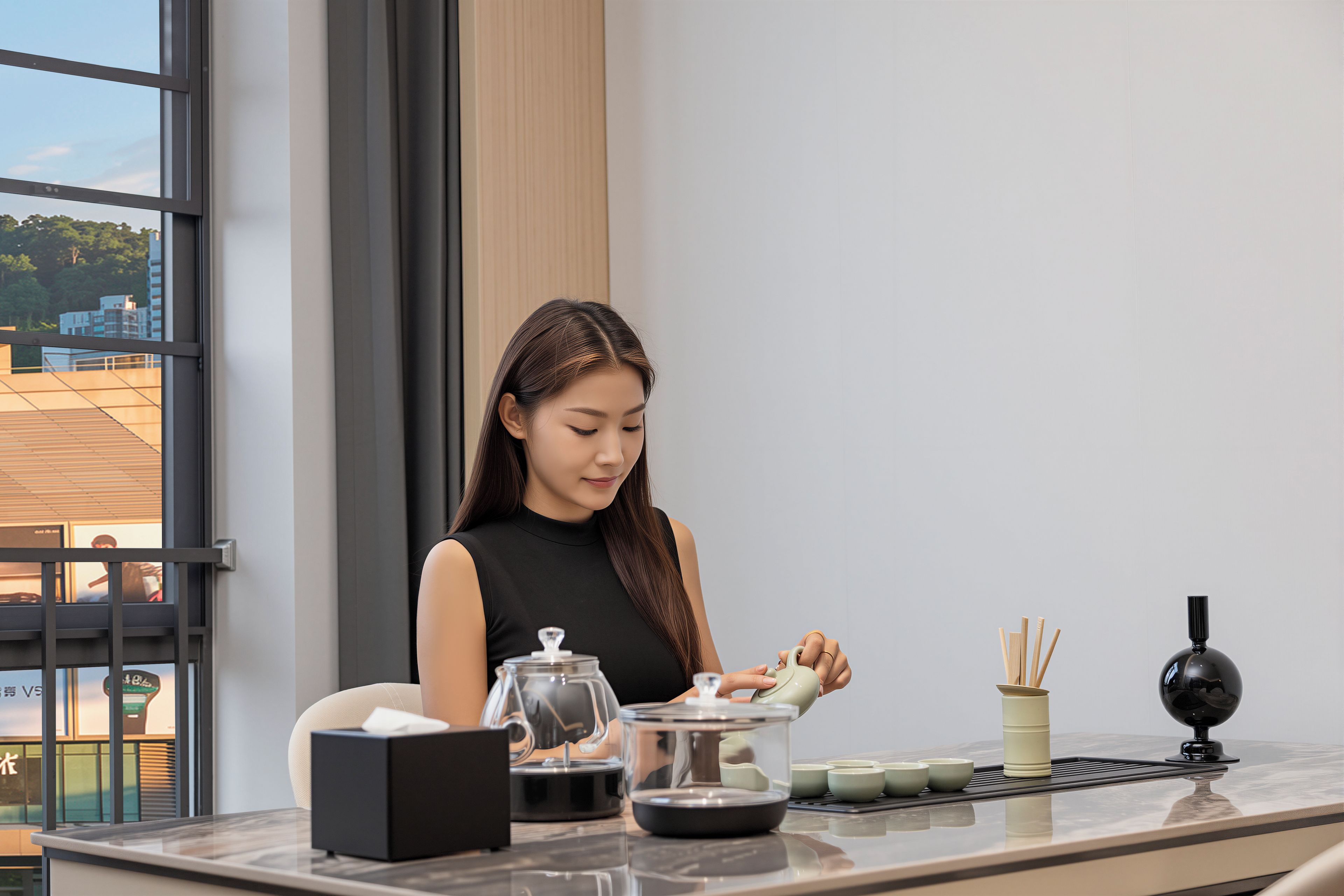 A woman seated at a table with a tea set, a black box, and a window with a view of buildings.