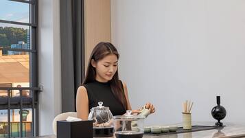 A woman seated at a table with a tea set, a black box, and a window with a view of buildings.