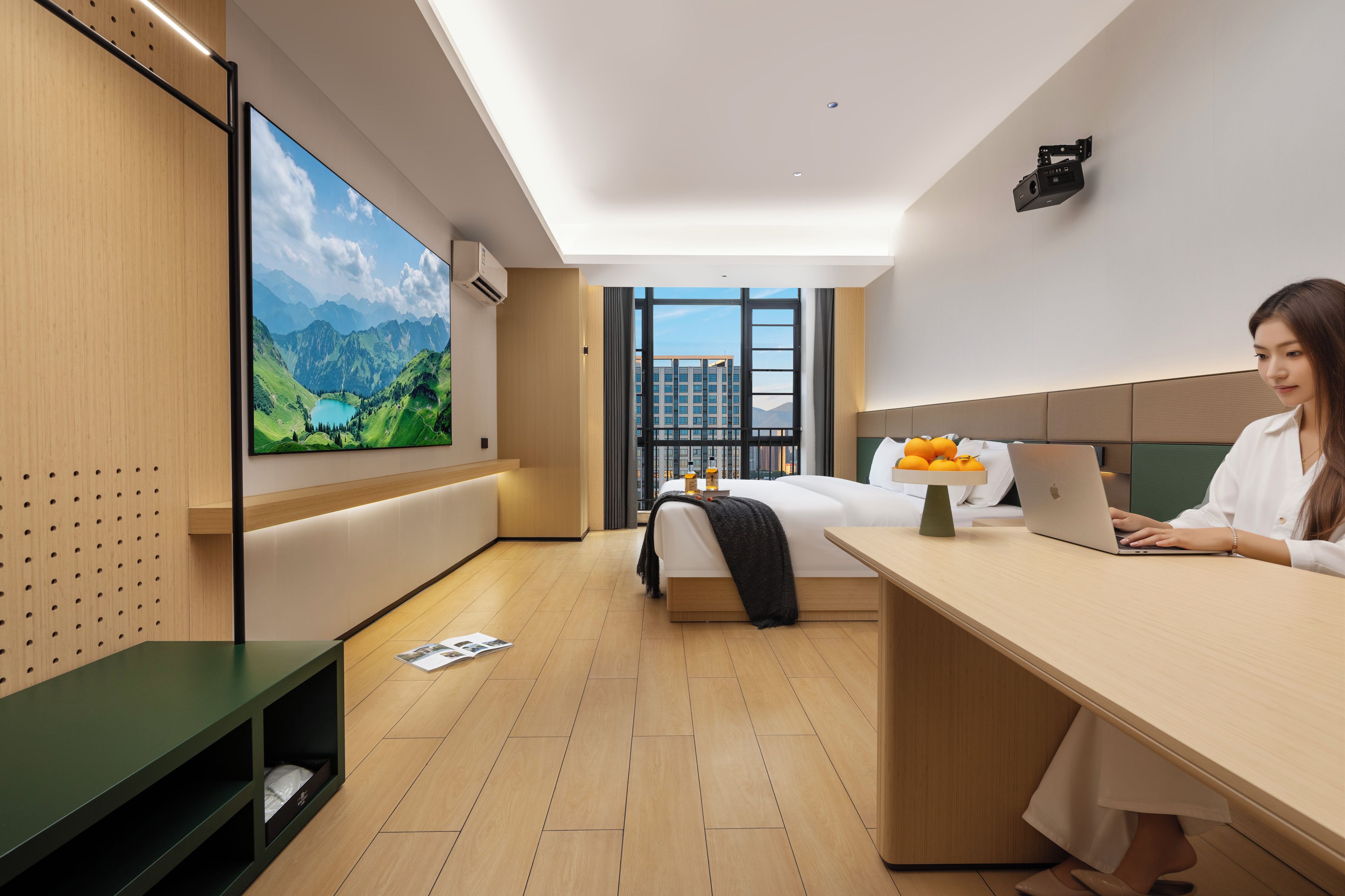 A modern hotel room with a large TV, a bed, a desk with a laptop, and a view of a cityscape through the window.