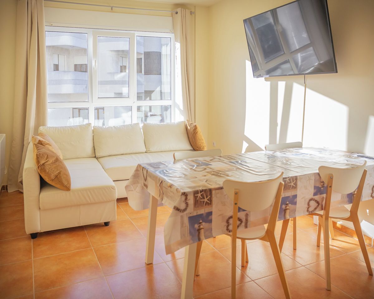 Una sala de estar con un sofá, una mesa de comedor con sillas, un televisor de pantalla plana montado en la pared y una ventana con cortinas.