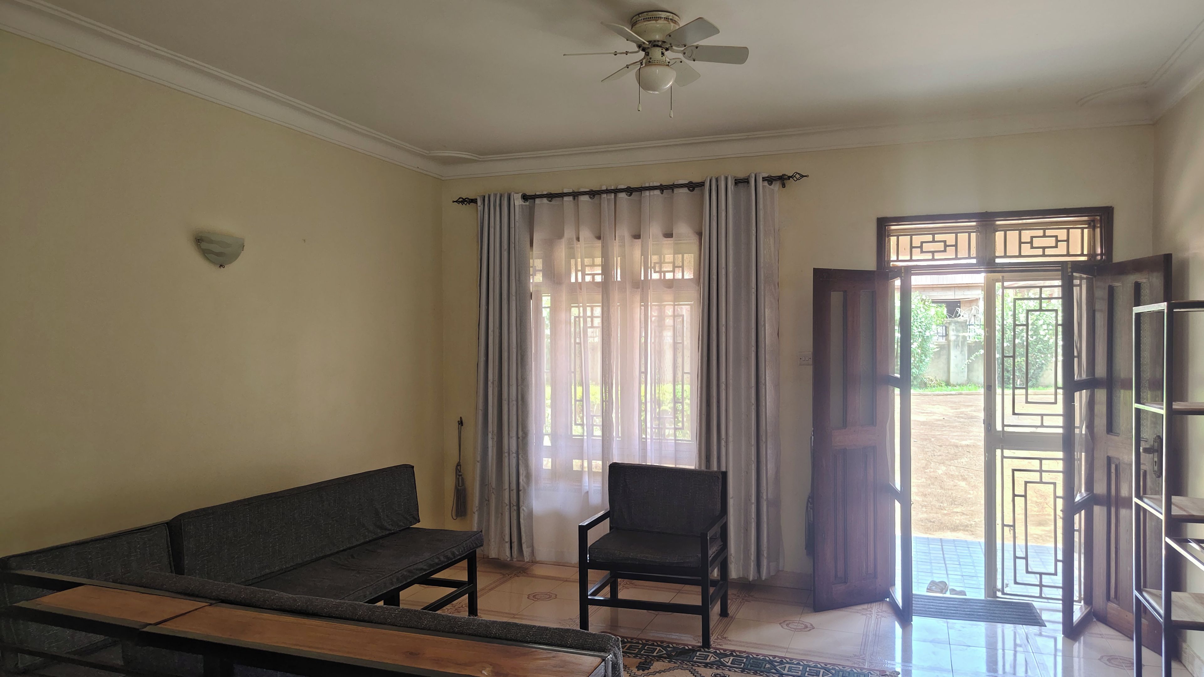 Une pièce comprenant un canapé, un fauteuil, un ventilateur de plafond, une porte ornée d’un motif géométrique et donnant sur l’extérieur.