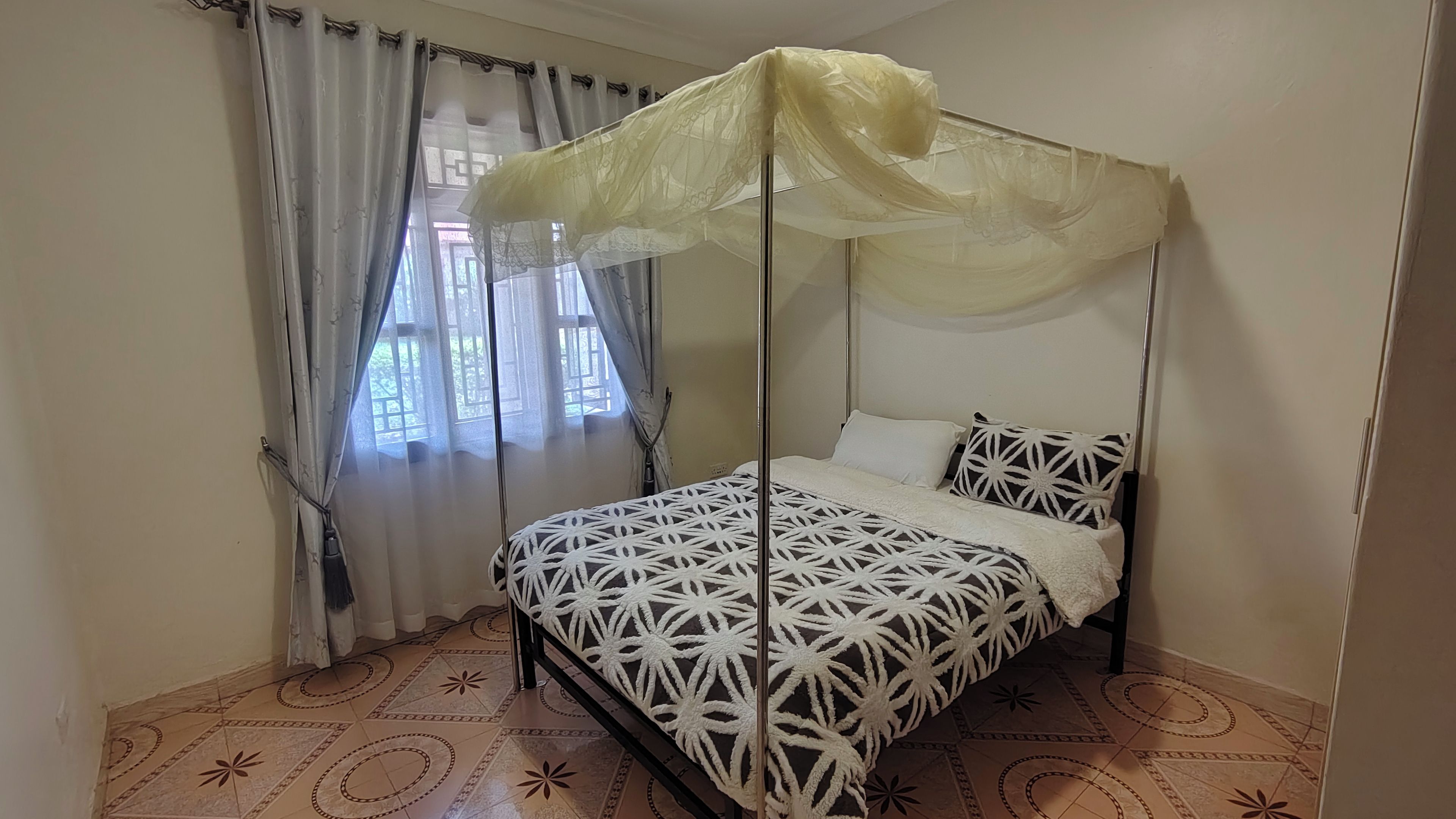 Un lit simple avec un baldaquin, une fenêtre avec des rideaux et des carreaux de sol à motifs.