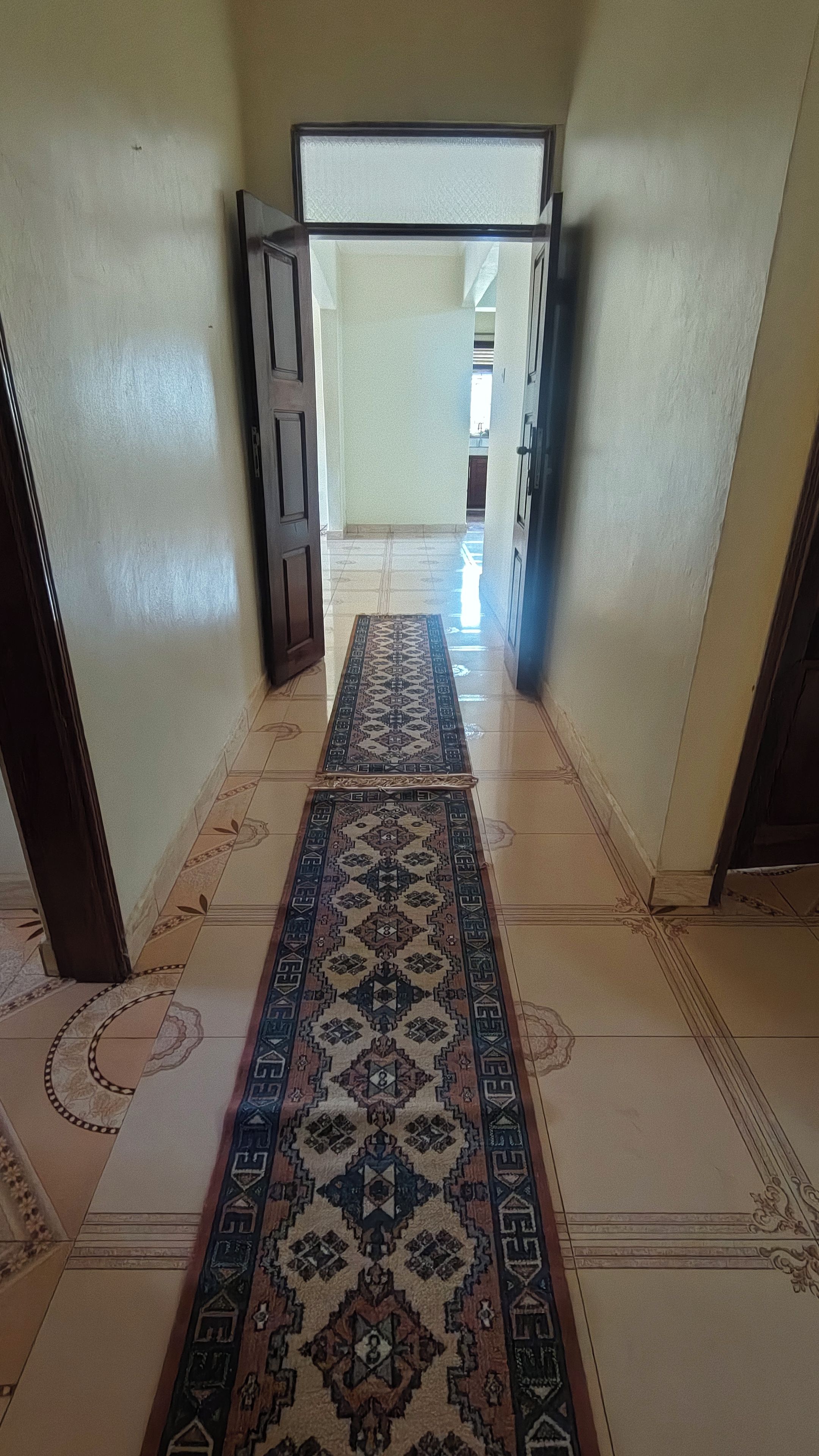 A hallway with a patterned carpet, wooden doors, and tiled flooring.