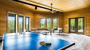 A ping pong table in a wooden room with large windows and a ceiling fan. - Milk & Honey 3BR Mountain Views | Indoor Pool, Hot Tub, Pool Table, Ping Pong (Sevierville)