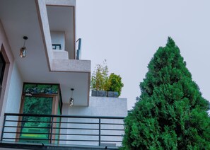 Un edificio moderno con un balcón, un ventanal grande y un árbol verde al frente.