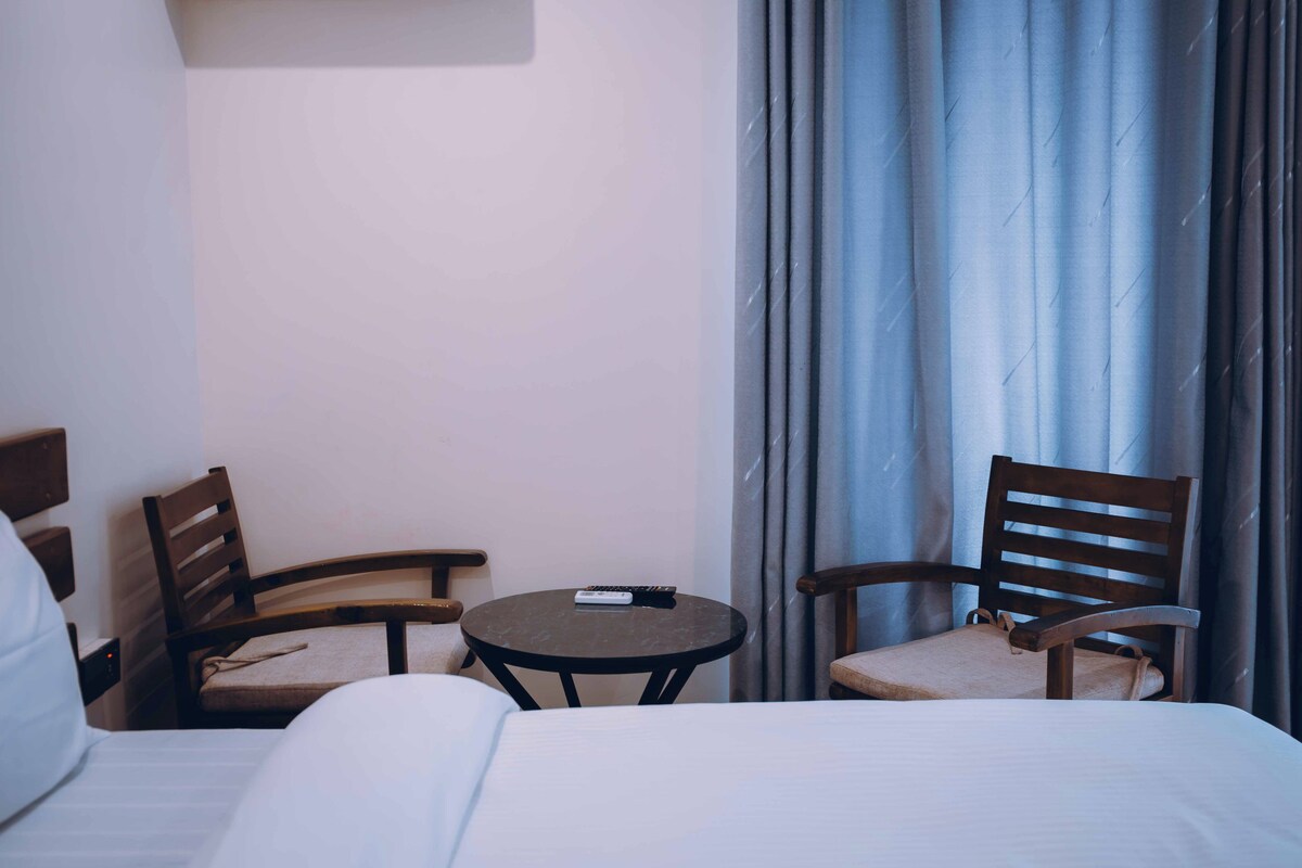 Habitación de hotel con cama, dos sillas de madera, una mesita redonda y un control remoto sobre la mesa.