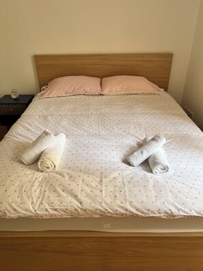 Een net opgemaakt bed met een geruit kussen, twee opgerolde handdoeken en een bedsprei met een patroon.