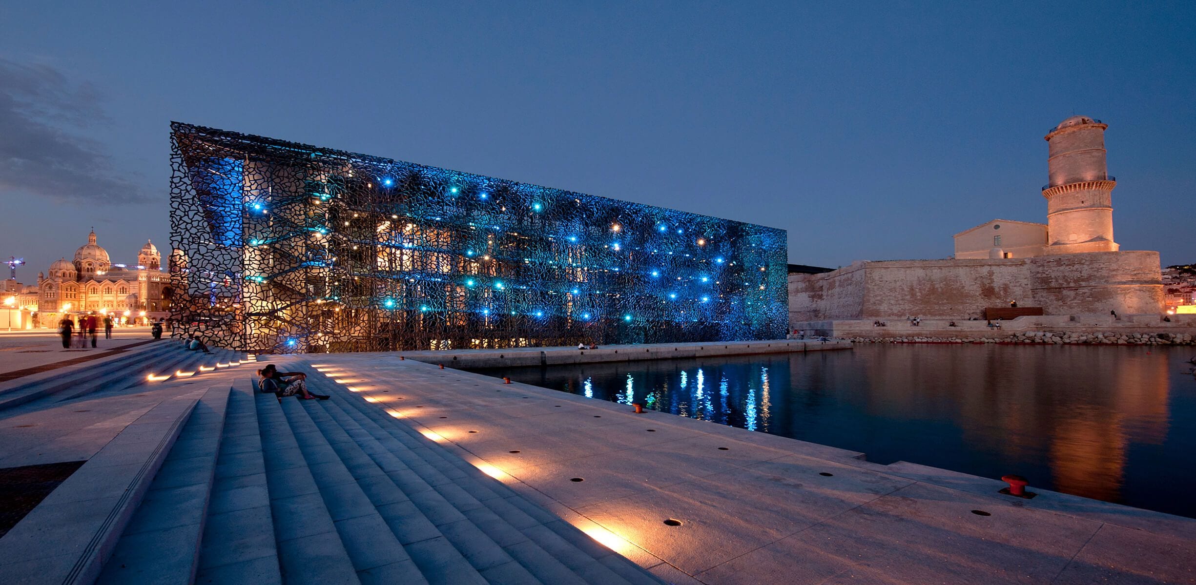 Een waterkantpromenade met een modern gebouw versierd met blauwe lichten, een rustig wateroppervlak en een historische toren.