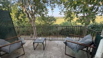 Een terras buiten met een tafel en twee stoelen, omgeven door een metalen hek, met uitzicht op een groen landschap.