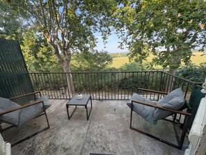 Une terrasse extérieure avec une table et deux chaises, entourée d’une rambarde en métal, donnant sur un paysage verdoyant.