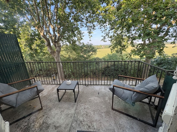 Eine Terrasse im Freien mit Tisch und zwei Stühlen, umgeben von einem Metallgeländer, mit Blick auf eine grüne Landschaft.