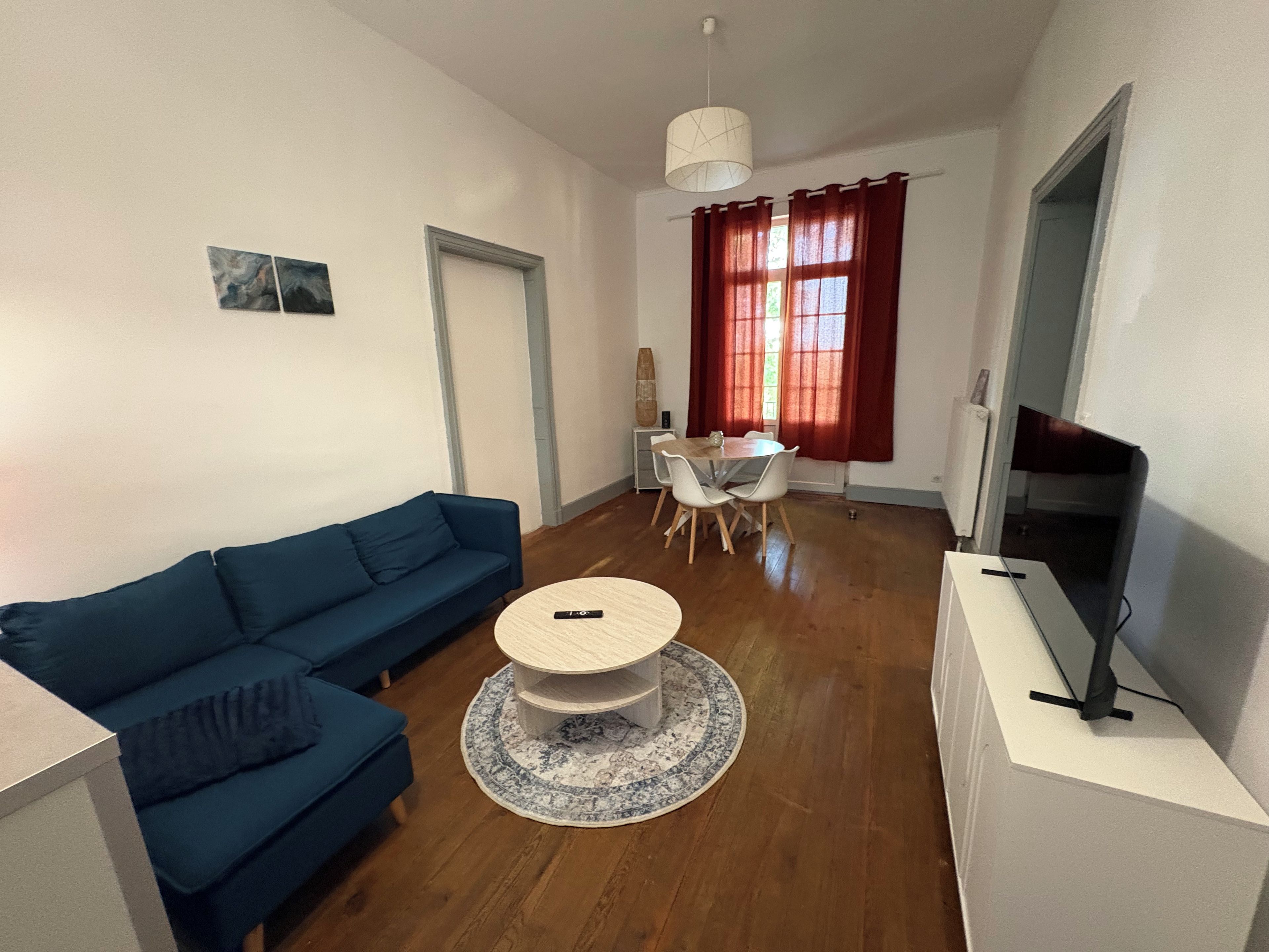 A living room with a blue sofa, a round coffee table, a dining table with chairs, a TV on a stand, and a window with curtains.