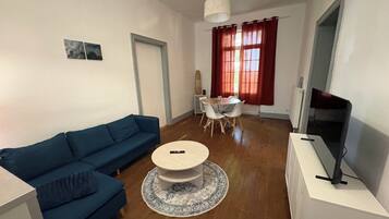 A living room with a blue sofa, a round coffee table, a dining table with chairs, a TV on a stand, and a window with curtains.