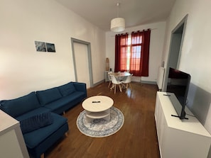 A living room with a blue sofa, a round coffee table, a dining table with chairs, a TV on a stand, and a window with curtains.