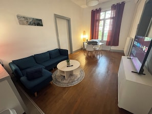 A living room with a blue sofa, a round coffee table, a dining table with chairs, a TV on a stand, and a window with curtains.