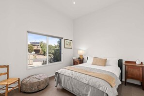 Ein Schlafzimmer mit einem großen Fenster, einem Bett mit grauer Bettdecke, einem Nachttisch mit Lampe, einem Holzbeistelltisch, einem Sessel und einem Sitzsack.