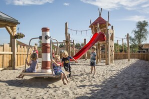 Un parque infantil con arena, un tobogán rojo y una estructura de madera para escalar.