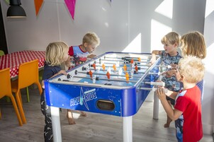 Un groupe d’enfants joue au baby-foot dans un espace intérieur bien éclairé, doté d’un plancher en bois et d’une table recouverte d’une nappe à carreaux rouges et blancs.