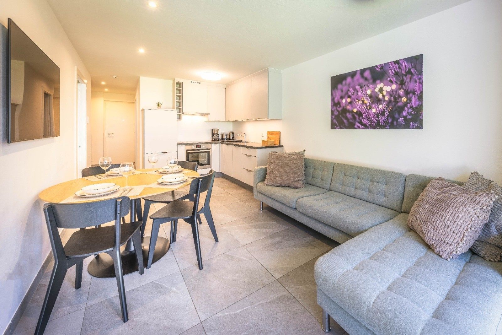 Ein modernes Wohnzimmer mit einem grauen Sofa, einem Esstisch für vier Personen und einer Küche im Hintergrund.