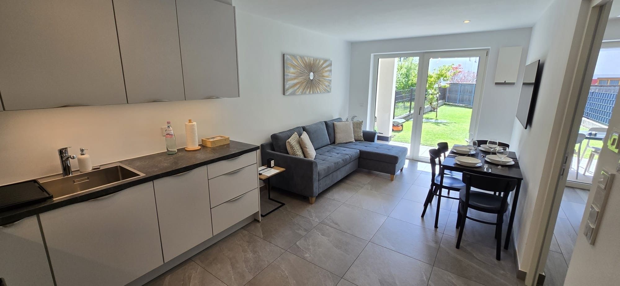 Et moderne køkken med en grå bordplade og hvide skabe, en grå sofa samt et spisebord med stole.