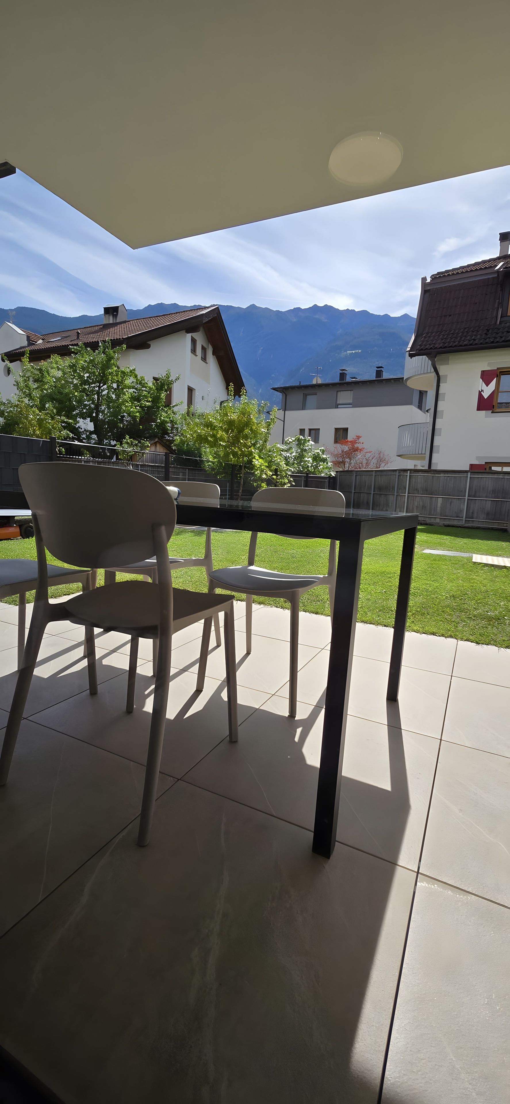 Eine Terrasse im Freien mit Tisch und Stühlen, Blick auf Berge und ein Gebäude mit rotem Fensterrahmen.