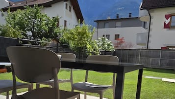 Eine Terrasse im Freien mit Tisch und Stühlen, Blick auf Berge und ein Gebäude mit rotem Fensterrahmen.