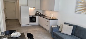 Una cucina compatta con frigorifero, forno e macchina da caffè. In zona soggiorno, un divano grigio con cuscini decorati.