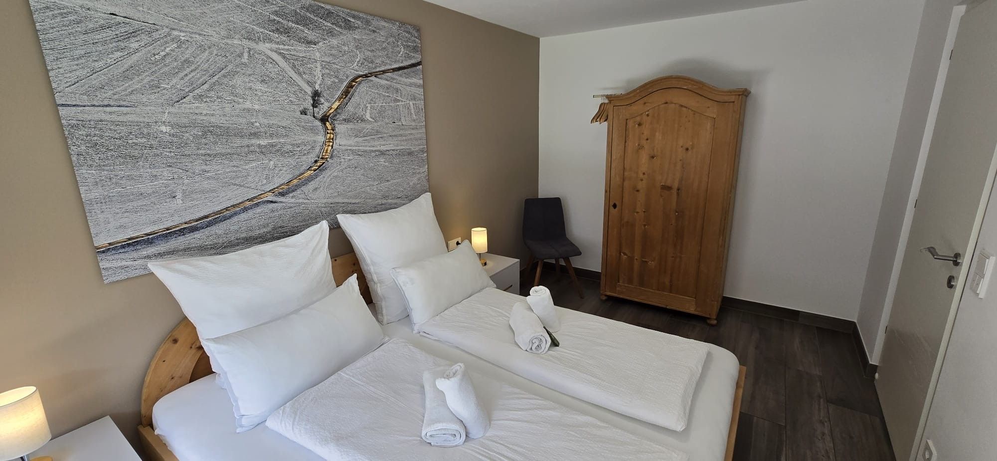 Ein Hotelzimmer mit einem Bett, Kissen, einem Holzschrank, einem Sessel und einem an der Wand hängenden Kunstwerk.