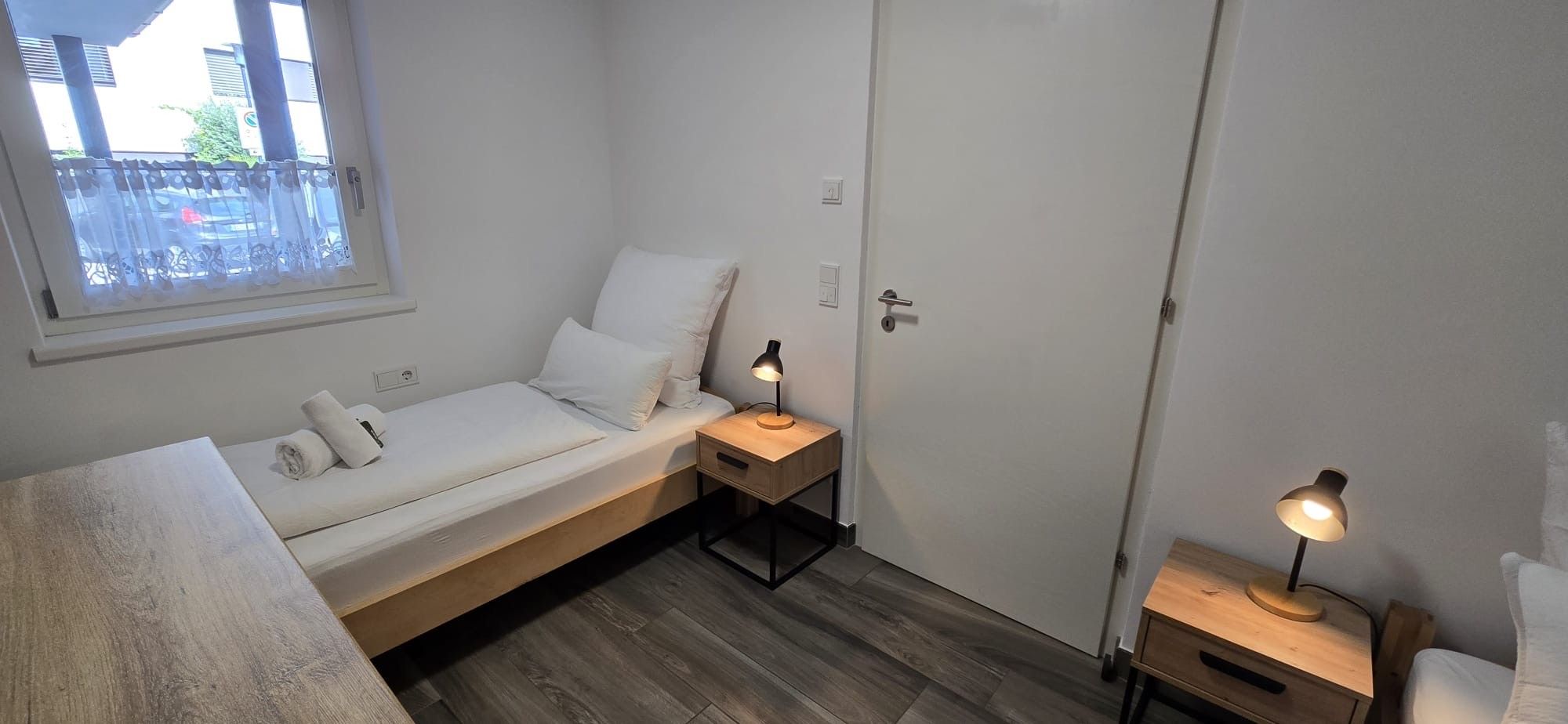 Una piccola camera con un letto singolo, una testiera in legno, un comodino con una lampada e una finestra vista esterna.