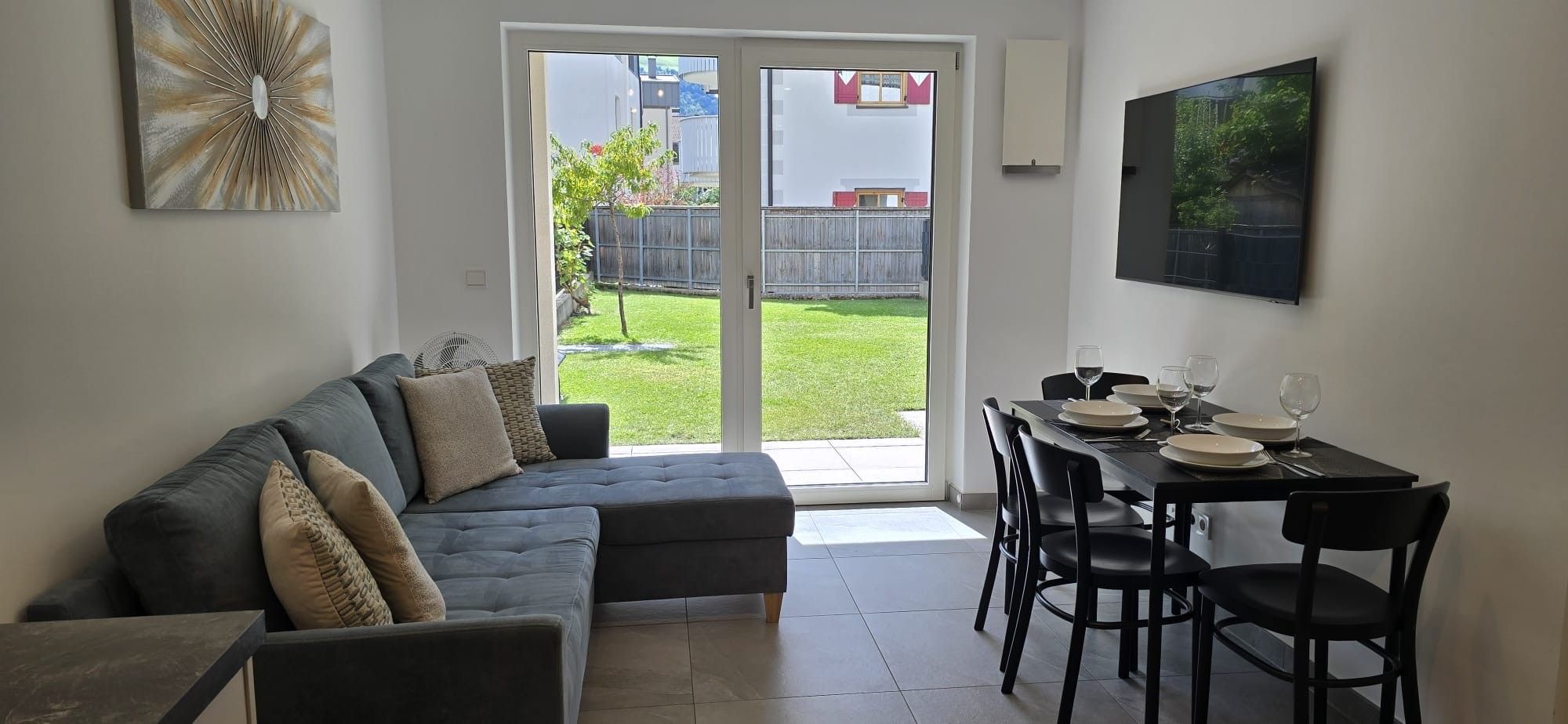 Ein modernes Wohnzimmer mit einem grauen Ecksofa, einem Esstisch für vier Personen und einer Schiebetür zu einem Garten.