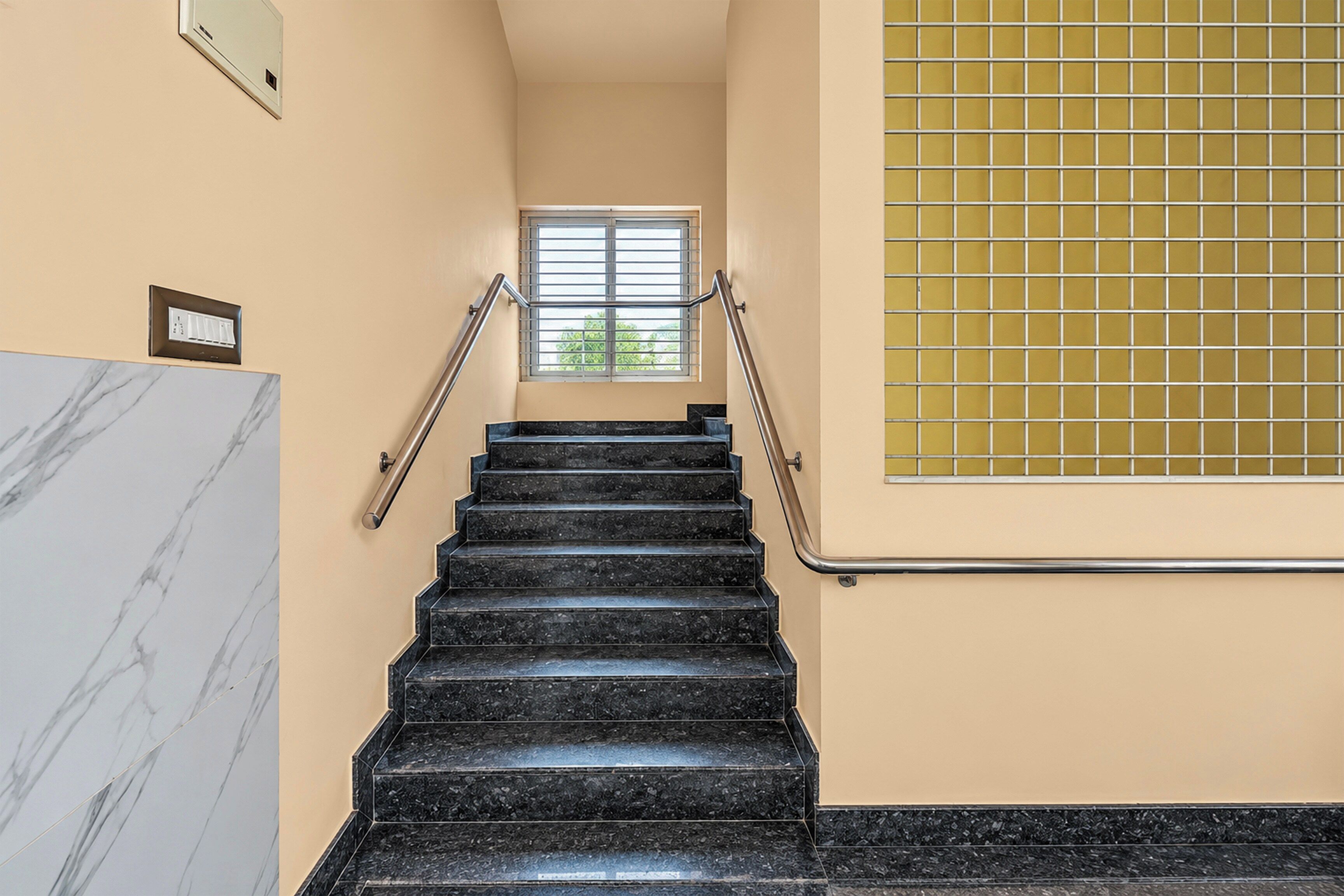 Una escalera con escalones de mármol negro, paredes color beige y una ventana con barrotes de metal.