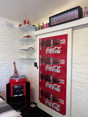 Ett rum med en vit tegelvägg, ett rött kylskåp med Coca-Cola-tema och hyllor med diverse föremål.