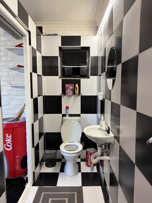 Ein kleines Badezimmer mit Toilette, Waschbecken und Spiegel. Ein Regal mit Toilettenartikeln und ein roter Coca-Cola-Kühler.