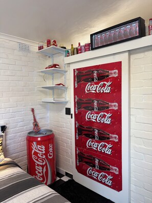 Ein Zimmer mit einer weißen Ziegelwand, einem Regal mit diversen Gegenständen, einem großen Coca-Cola-Poster und einer Coca-Cola-Dose.