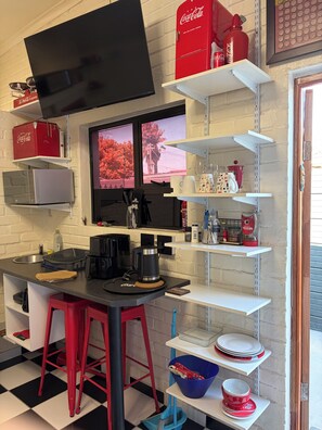 Ett kök med en svart bänkskiva, röda barstolar, en kaffemaskin och hyllor med diverse föremål, inklusive en kylbox för Coca-Cola.