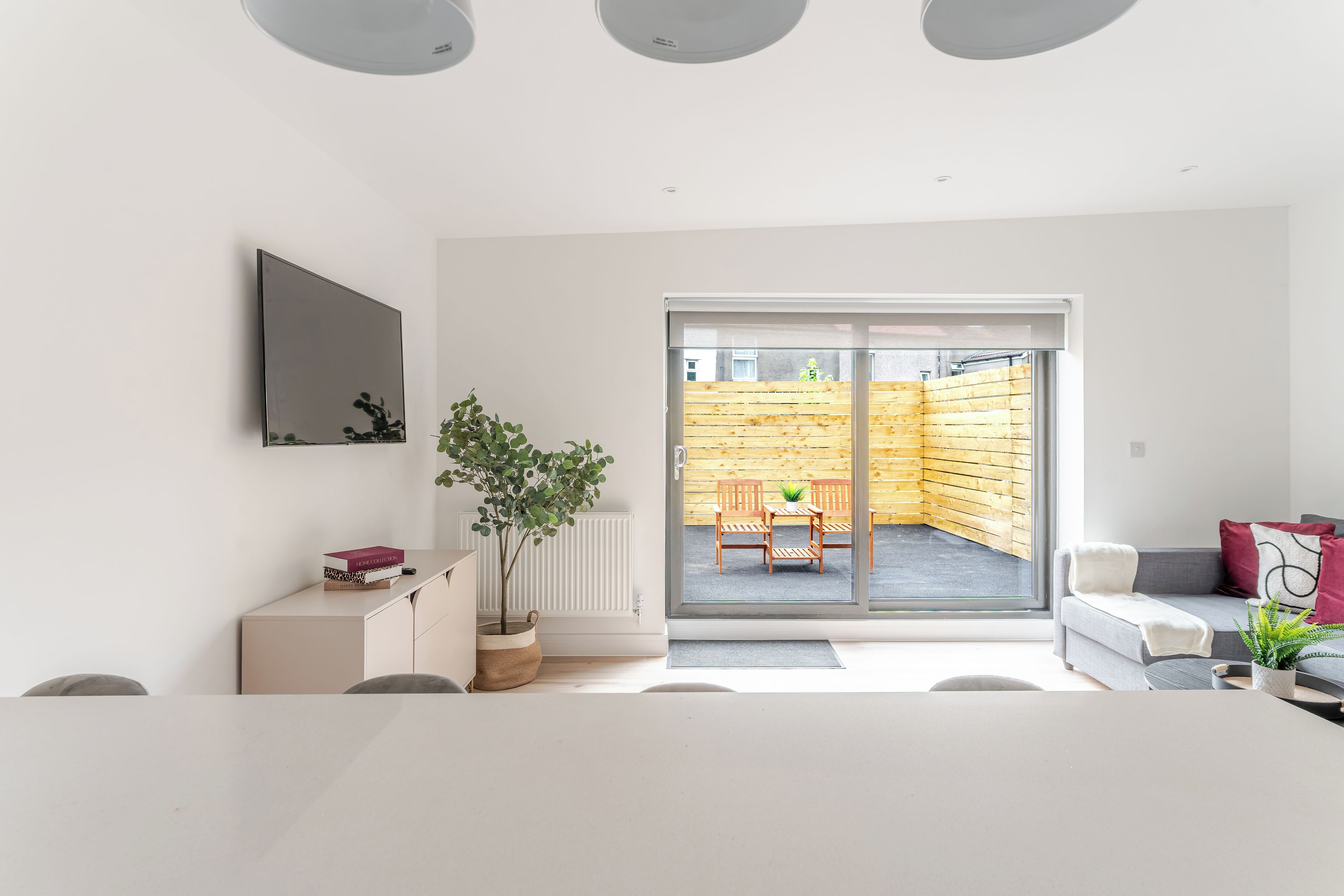 Un moderno salón con un televisor de pantalla plana, un sofá, una mesa de centro y un ventanal corredizo que da a un área exterior con una valla de madera y un banco.
