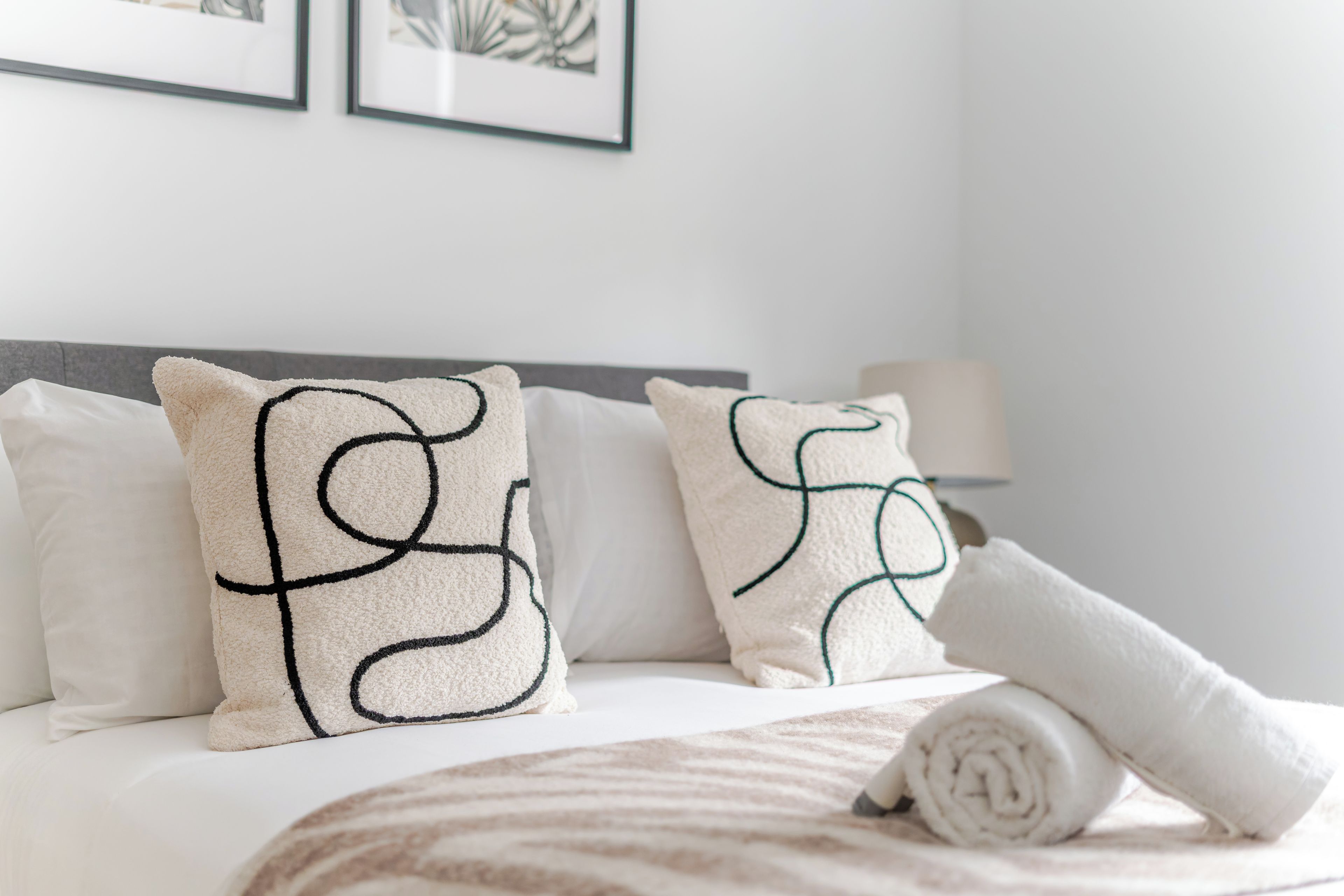 Cama bem arrumada com roupa de cama branca e manta listrada, dois travesseiros com bordado preto e uma toalha enrolada.