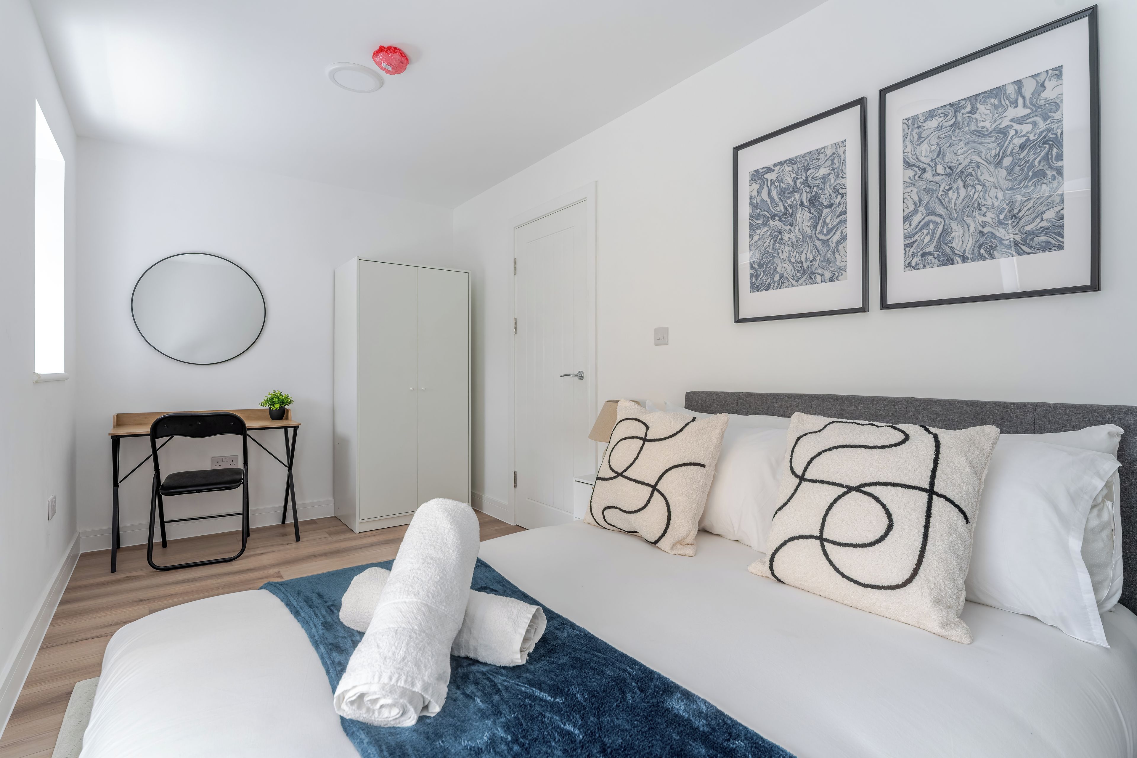 A bedroom with a bed, a desk, a chair, a wardrobe, and two framed artworks on the wall.
