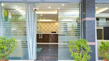 Entrada de hotel con puerta de vidrio, mostrador de recepción y plantas en macetas.