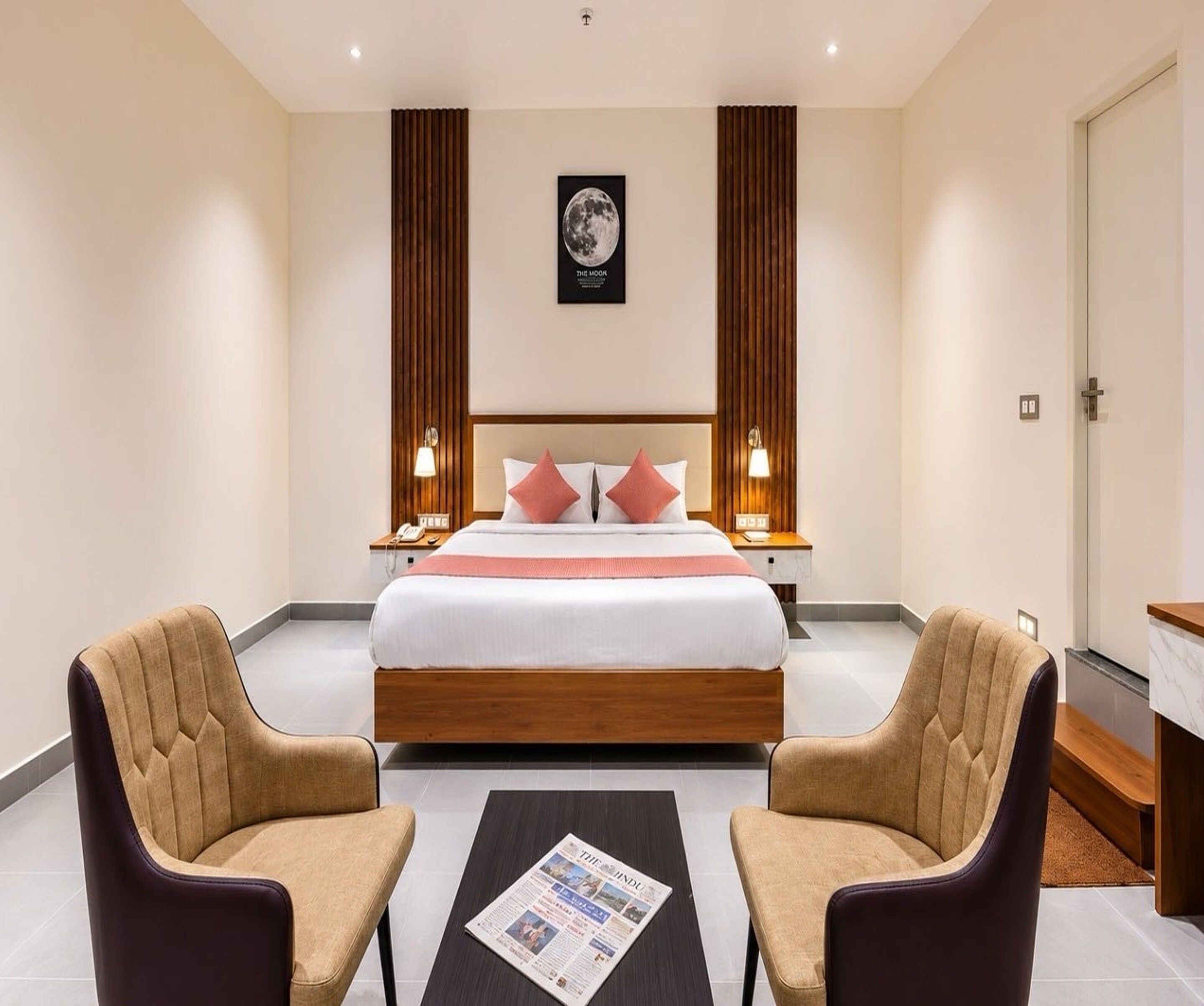 A hotel room with a large bed, two bedside tables, a nightstand, a desk, two armchairs, and a coffee table with a newspaper.