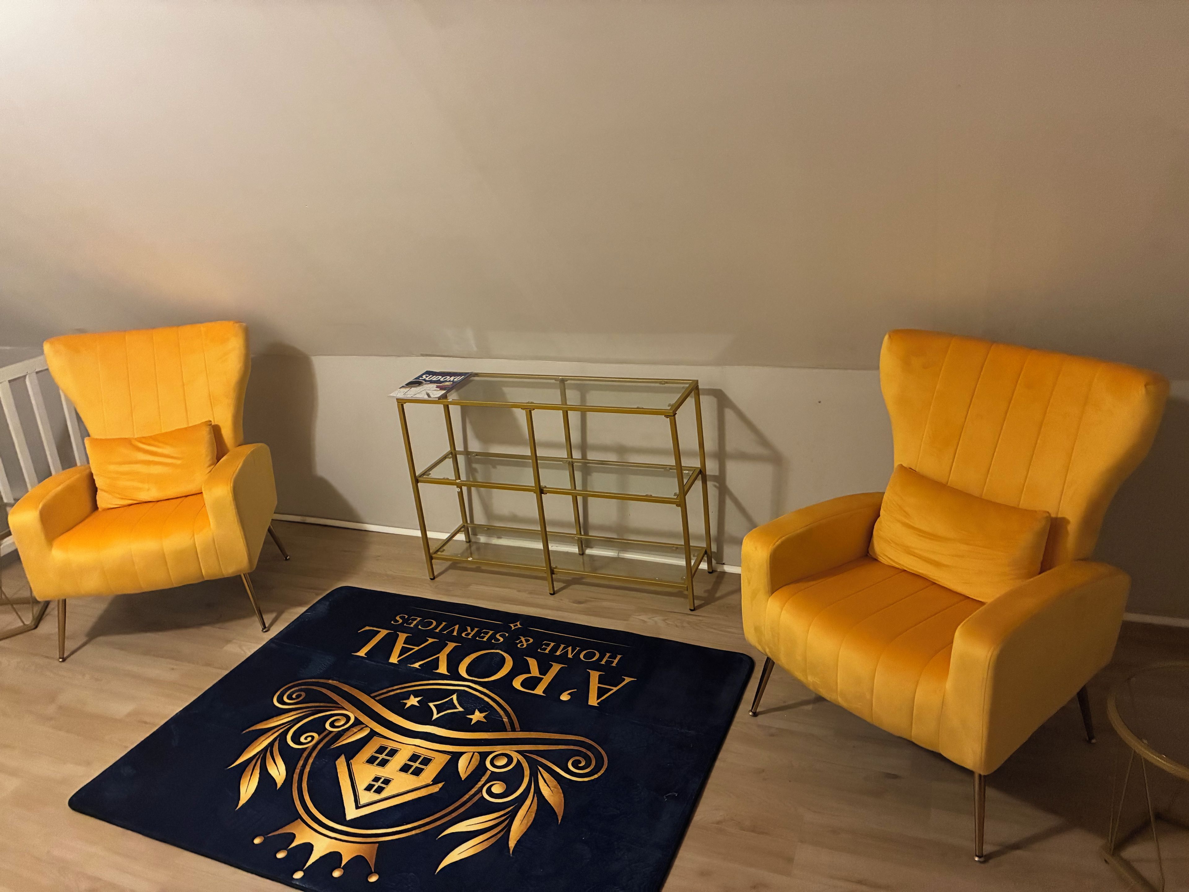 A room with two yellow armchairs, a gold-framed side table, and a dark blue rug with a crest design.