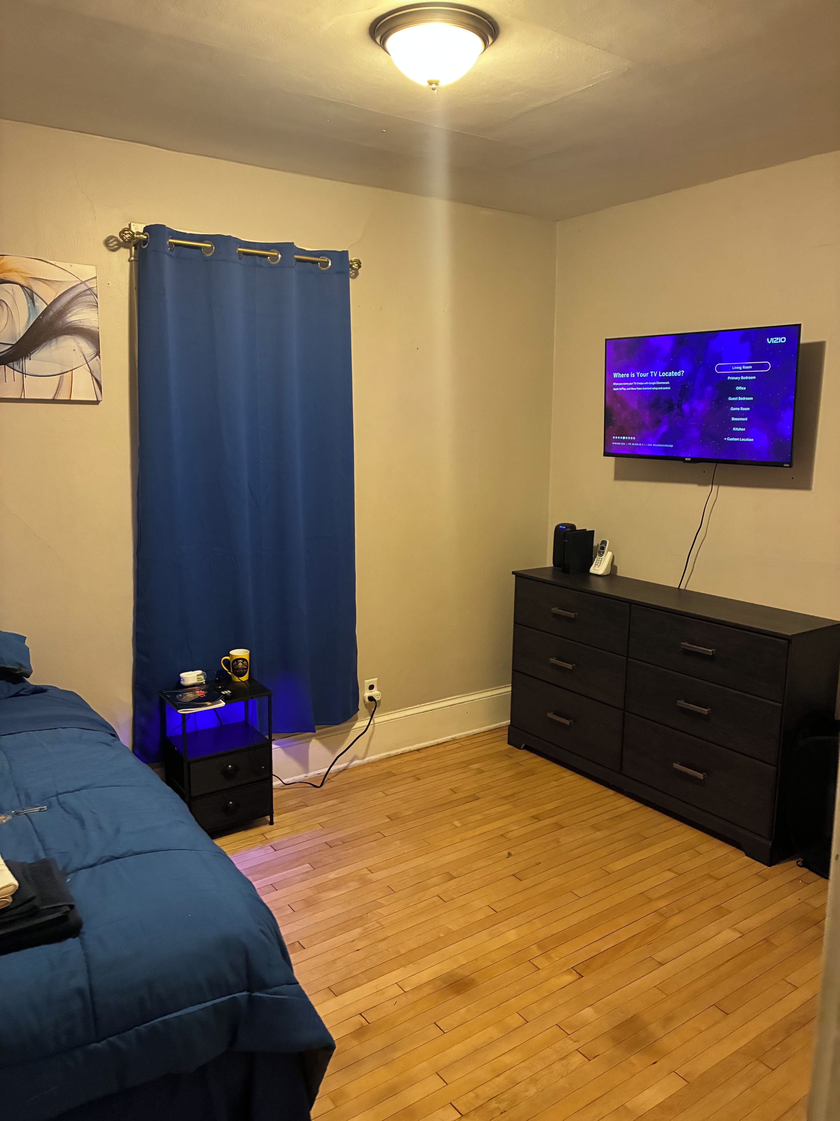 A bedroom with a bed, a dresser, a TV on the wall, and a nightstand with a lamp.