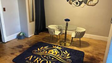 A room with a glass table and two chairs, a dark blue area rug with a logo, a wall-mounted abstract art piece, and a dark curtain.
