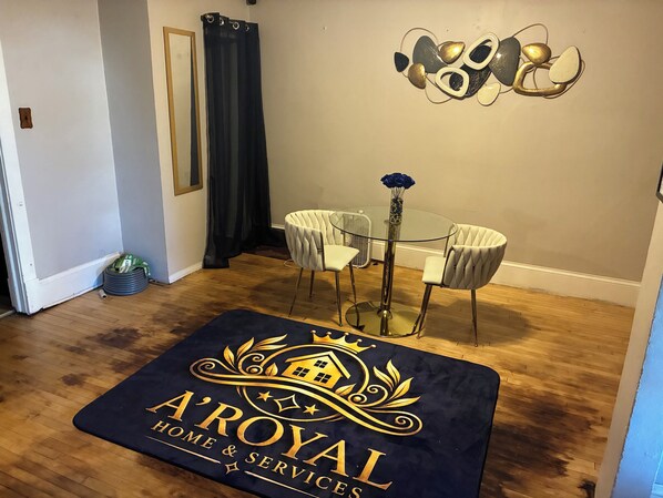 A room with a glass table and two chairs, a dark blue area rug with a logo, a wall-mounted abstract art piece, and a dark curtain.