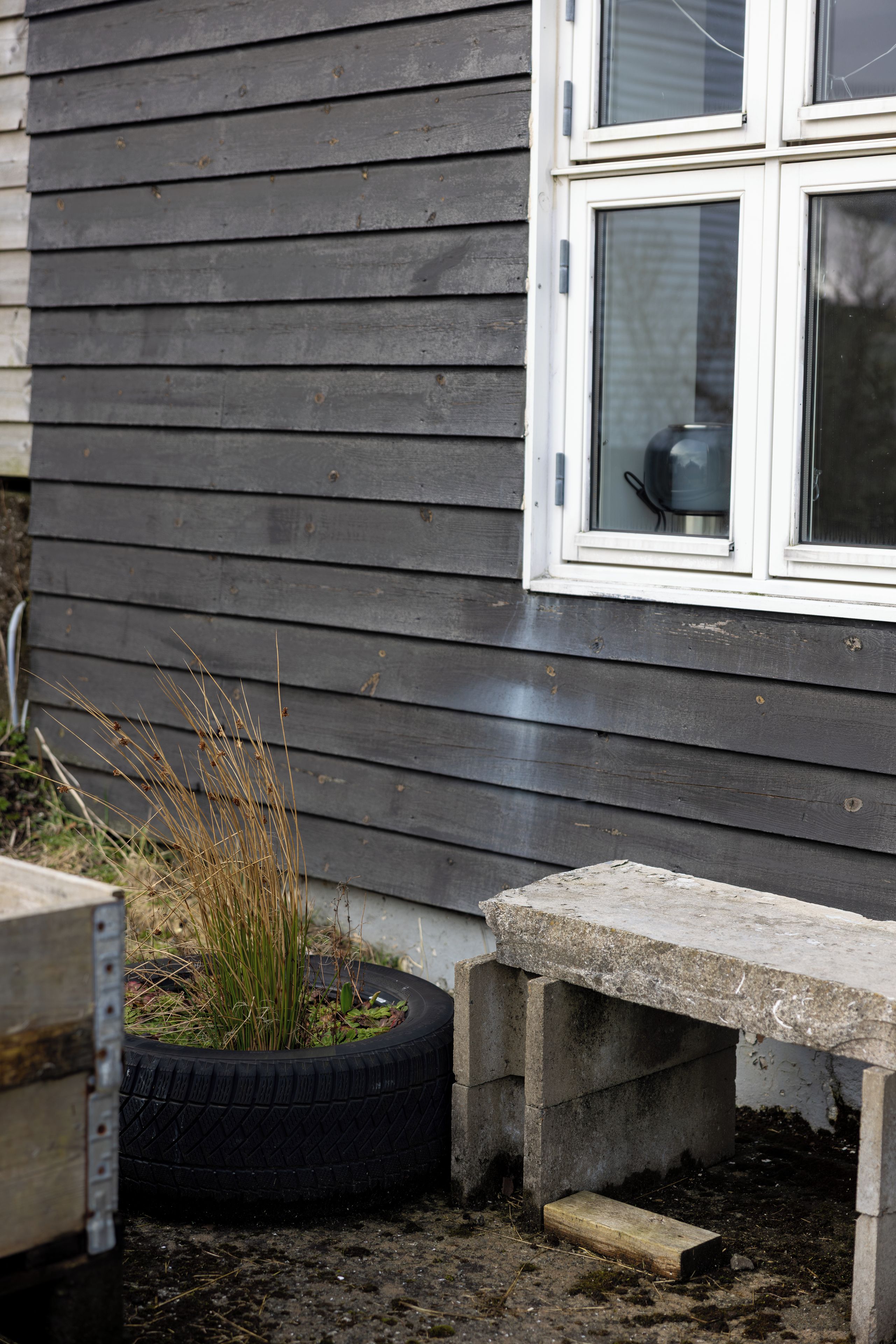 Een betonnen bank en een bandenplanter met planten, naast een huis met donkere bekleding en een witte raamkozijn.